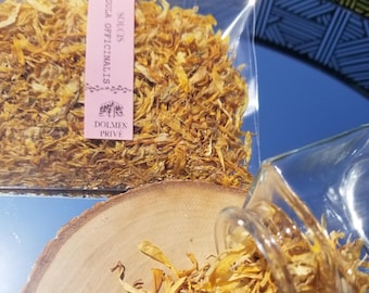 Może przedstawiać: Przezroczysty szklany słoik i plastikowa torba wypełniona suszonymi, żółtymi płatkami nagietka. Torba ma różową etykietę z tekstem i logo. Niektóre płatki są rozrzucone na drewnianym plastrze. Tło to powierzchnia odbijająca światło.
