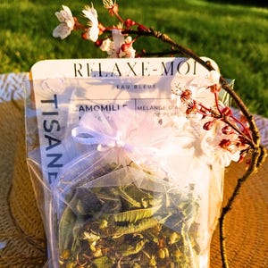 Puede incluir: Una bolsa blanca con una cinta blanca y una etiqueta que dice "RELAX-MOI EAU BLEUE CAMOMILLE MELANGE D'AL DE PLANTES TISANE". La bolsa está llena de hierbas secas y está sentada en un sombrero de paja.