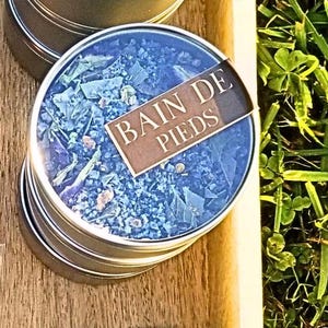 Peut inclure: Une boîte en argent contenant un mélange bleu et blanc. La boîte porte une étiquette brune sur laquelle est inscrit "Bain de Pieds".