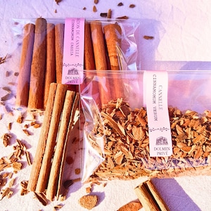 Peut inclure: Gros plan sur des bâtons et morceaux de cannelle, certains dans des sachets en plastique transparent avec des étiquettes roses. Les étiquettes indiquent "Bâton de Cannelle" et "Cinnamonum Cassia" et "Cinnamonum Verum". La cannelle est de couleur marron chaud, sur un fond clair.