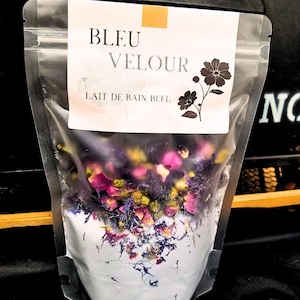 Peut inclure: Un sac en plastique transparent rempli d'un bain moussant blanc, de pétales de fleurs bleues et violettes et de feuilles vertes. Le sac porte une étiquette blanche avec le texte "BLEU VELOUR LAIT DE BAIN BLEU" et une illustration de fleur noire.