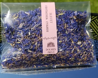 Może przedstawiać: Przezroczysty pakiet wypełniony suszonymi płatkami chabra bławatka. Różowa etykieta zawiera napis "BLEUET Centaurea cyanus" i "DOLMEN PRIVÉ" z małą ilustracją. Opakowanie leży na odblaskowej powierzchni, z zieloną trawą w tle.