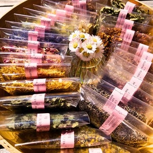 Puede incluir: Una bandeja de madera llena de pequeñas bolsas transparentes que contienen varias hierbas y flores secas. Cada bolsa está sellada con una etiqueta de cinta rosa. Un pequeño jarrón de margaritas está en el centro.