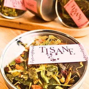 Peut inclure: Gros plan sur plusieurs boîtes métalliques remplies d'herbes et de fleurs séchées. Chaque boîte porte une étiquette rose avec le mot "TISANE" en lettres violet foncé. Les boîtes sont disposées sur une surface en bois clair, suggérant un produit à base de tisane.