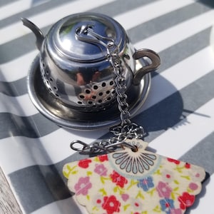 Può includere: Un infusore per tè in argento a forma di teiera con una catena e un ciondolo a ventaglio in tessuto con motivo floreale.