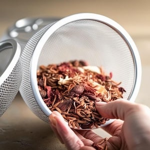 May include: A silver mesh tea infuser with a lid, filled with dried herbs and flowers. The infuser is being held by a hand.