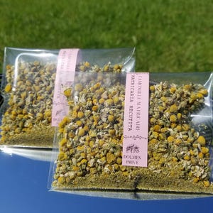 Può includere: Due sacchetti trasparenti pieni di fiori e foglie di camomilla essiccati. Ogni sacchetto ha un'etichetta rosa con testo in lingua straniera. I sacchetti sono su una superficie riflettente con uno sfondo erboso verde.