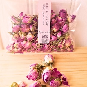 Könnte beinhalten: Ein klarer Beutel gefüllt mit getrockneten rosa Rosenknospen und Blütenblättern. Das Etikett trägt die Aufschrift "Bouton de Rose Rosa Damascena" mit dem Logo "Dolmen Privé". Einige Rosenknospen liegen auf einer hellen Holzoberfläche.