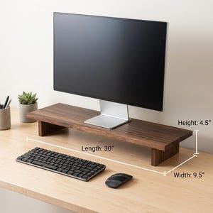 May include: A dark brown wooden monitor stand with a computer monitor on top. The stand is 30 inches long, 9.5 inches wide, and 4.5 inches high. A black keyboard and mouse are in front of the stand. The desk is light brown.