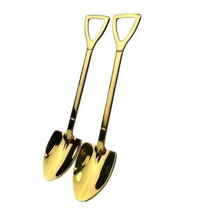 May include: Two gold-colored metal spoons shaped like miniature shovels. The spoons are arranged side-by-side with the handles pointing upwards.