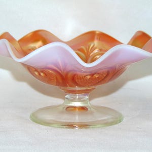 May include: A vintage iridescent glass bowl with a scalloped edge and a footed base. The bowl is a peachy-orange color with a white iridescent finish.