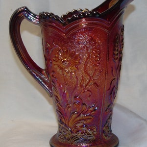 May include: A red iridescent glass pitcher with a floral design. The pitcher has a scalloped rim and a flared base.