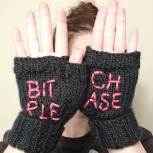 May include: Dark gray knitted fingerless gloves with the words "BIT" and "PLE" embroidered in pink on one glove and "CHASE" and "ASE" on the other. The gloves are made of a thick yarn.