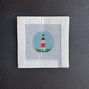 May include: A small, square needlepoint canvas featuring a lighthouse design. The lighthouse is red, white, and black, set against a blue sky and green grass. The canvas is surrounded by a white border and is on a black background.