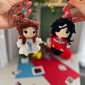 May include: Two handmade crochet doll keyrings. One doll has brown hair and a white dress, the other has black hair and a red outfit. Both dolls have beaded accents and are attached to silver keyrings.