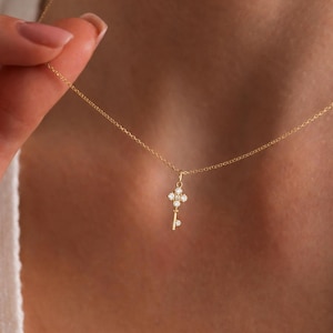 May include: A delicate gold necklace featuring a key pendant adorned with small, sparkling gemstones. The key's head has a four-petal design. The necklace is held up against a person's neck, showcasing the jewelry's fine details and elegant simplicity.