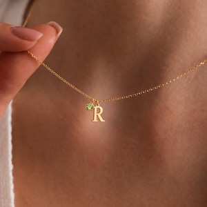 Peut inclure: Un délicat collier en or avec un pendentif en forme de lettre "R" et un petit charme de pierre précieuse verte. Le collier est présenté sur un fond de teint, mettant en valeur le design et la couleur du bijou.