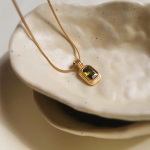 Può includere: Una collana color oro con un ciondolo rettangolare caratterizzato da una gemma sfaccettata color verde oliva. Il ciondolo è sospeso a una sottile e flessibile catena a serpente color oro, con una cornice in metallo lucido e dettagli a strati.