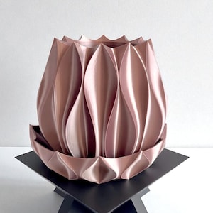 May include: A decorative, petal-shaped vase in a metallic rose gold color. The vase sits on a black, geometric stand. The vase has a textured, layered design, and the stand has a minimalist, modern aesthetic. The background is white.