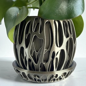 May include: A decorative metallic silver plant pot with a matching saucer. The pot has a unique openwork design, with a pattern of curved lines and shapes. The pot holds a green leafy plant. The pot and saucer are sitting on a white surface.