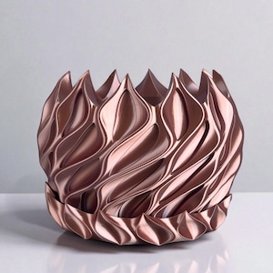 A 3D‑printed planter with a curvy cylindrical form, featuring bold vertical chunky wavy ridges that twist elegantly from top to bottom. The pot includes drainage holes and comes with a matching drip tray that mirrors the same sculptural surface design. Made from glossy Rose Gold Silk PLA, the planter has a luminous sheen that enhances its contemporary art aesthetic.