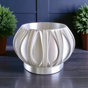 Pode incluir: Um vaso decorativo branco com um design texturizado único. O vaso tem uma forma arredondada com painéis verticais em forma de gota e detalhes estampados. Assenta numa base circular e é adequado para plantas de interior.