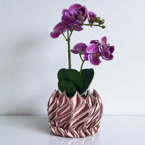 A 3D‑printed planter with a curvy cylindrical form, featuring bold vertical chunky wavy ridges that twist elegantly from top to bottom. The pot includes drainage holes and comes with a matching drip tray that mirrors the same sculptural surface design. Made from glossy Rose Gold Silk PLA, the planter has a luminous sheen that enhances its contemporary art aesthetic.