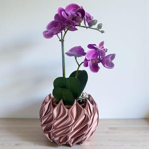 A 3D‑printed planter with a curvy cylindrical form, featuring bold vertical chunky wavy ridges that twist elegantly from top to bottom. The pot includes drainage holes and comes with a matching drip tray that mirrors the same sculptural surface design. Made from glossy Rose Gold Silk PLA, the planter has a luminous sheen that enhances its contemporary art aesthetic.