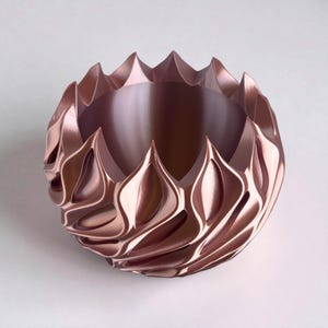 A 3D‑printed planter with a curvy cylindrical form, featuring bold vertical chunky wavy ridges that twist elegantly from top to bottom. The pot includes drainage holes and comes with a matching drip tray that mirrors the same sculptural surface design. Made from glossy Rose Gold Silk PLA, the planter has a luminous sheen that enhances its contemporary art aesthetic.