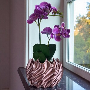 A 3D‑printed planter with a curvy cylindrical form, featuring bold vertical chunky wavy ridges that twist elegantly from top to bottom. The pot includes drainage holes and comes with a matching drip tray that mirrors the same sculptural surface design. Made from glossy Rose Gold Silk PLA, the planter has a luminous sheen that enhances its contemporary art aesthetic.