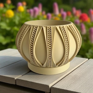 May include: A beige, decorative planter with an intricate, geometric design. The planter has a rounded shape with vertical, teardrop-shaped cutouts and a decorative band around the middle. It sits on a circular base and is placed on a wooden surface.
