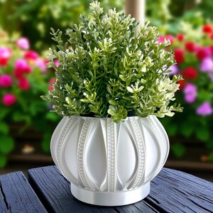 May include: A white, decorative planter with a textured, geometric design, holding a vibrant green plant. The planter has a round shape with vertical, curved lines and a circular base. The plant has small, green leaves. The background is blurred with colorful flowers.
