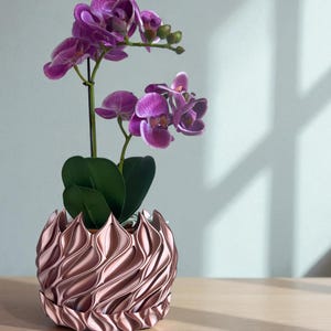 A 3D‑printed planter with a curvy cylindrical form, featuring bold vertical chunky wavy ridges that twist elegantly from top to bottom. The pot includes drainage holes and comes with a matching drip tray that mirrors the same sculptural surface design. Made from glossy Rose Gold Silk PLA, the planter has a luminous sheen that enhances its contemporary art aesthetic.