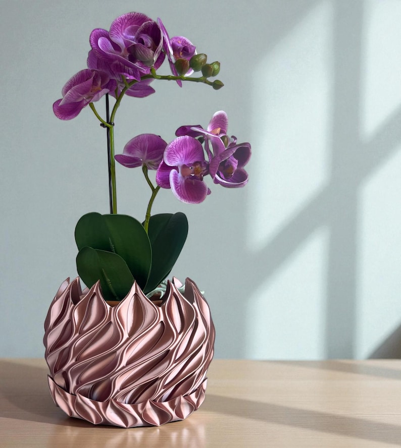 A 3D‑printed planter with a curvy cylindrical form, featuring bold vertical chunky wavy ridges that twist elegantly from top to bottom. The pot includes drainage holes and comes with a matching drip tray that mirrors the same sculptural surface design. Made from glossy Rose Gold Silk PLA, the planter has a luminous sheen that enhances its contemporary art aesthetic.