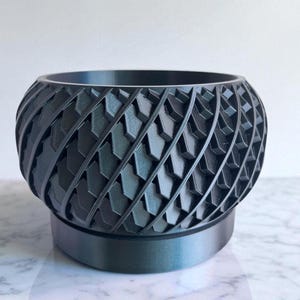 Puede incluir: Una maceta gris oscuro, impresa en 3D, con un patrón geométrico. La maceta tiene una forma redondeada con una superficie texturizada y se asienta sobre una base a juego. El diseño presenta formas de flecha repetidas, creando un efecto espiral.
