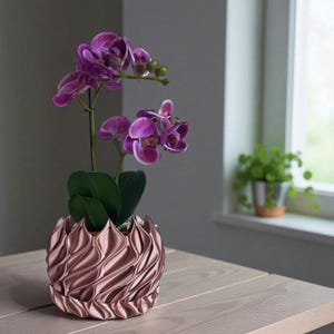 A 3D‑printed planter with a curvy cylindrical form, featuring bold vertical chunky wavy ridges that twist elegantly from top to bottom. The pot includes drainage holes and comes with a matching drip tray that mirrors the same sculptural surface design. Made from glossy Rose Gold Silk PLA, the planter has a luminous sheen that enhances its contemporary art aesthetic.