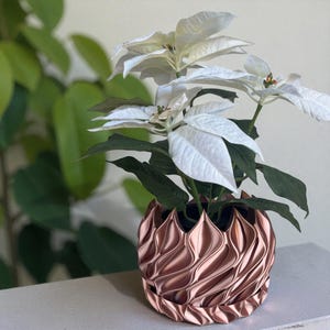A 3D‑printed planter with a curvy cylindrical form, featuring bold vertical chunky wavy ridges that twist elegantly from top to bottom. The pot includes drainage holes and comes with a matching drip tray that mirrors the same sculptural surface design. Made from glossy Rose Gold Silk PLA, the planter has a luminous sheen that enhances its contemporary art aesthetic.