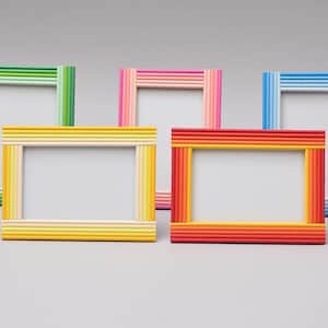 May include: Five colourful picture frames in shades of green, pink, yellow, red, and blue. Each frame has a rectangular opening and is constructed with multiple parallel rods, creating a striped effect. The frames are arranged on a light grey surface.
