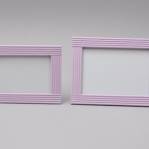 May include: Two rectangular picture frames in a soft lavender colour. The frames are constructed with horizontal and vertical rods, creating a textured, linear design. The frames are empty, ready to display photos or artwork. The frames are of different sizes.