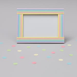 May include: A rectangular picture frame in pastel shades of pink, yellow, blue, and lavender. The frame is decorated with vertical and horizontal stripes. Confetti in matching colours is scattered around the frame on a grey surface.