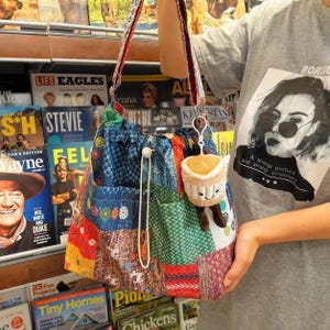 May include: A patchwork tote bag featuring a variety of colorful fabric patterns in blue, green, red, and orange. The bag has a drawstring closure with a white cord and a small, plush, smiling coffee cup charm attached to the strap.