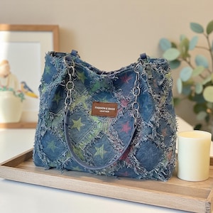 May include: A denim tote bag with a patchwork design featuring star patterns in various colors. The bag has chain handles and a leather tag that reads "FASHION & BAGS LEATHER". The bag is on a wooden tray.