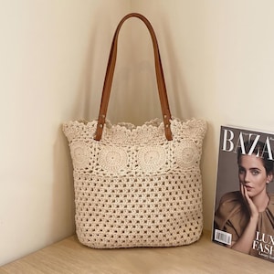 May include: A cream-colored crochet tote bag with brown leather handles. The bag features a decorative scalloped edge and circular patterns along the top. A magazine with the word "BAZAAR" is visible on the right.