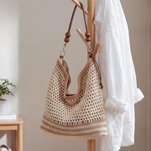 Könnte beinhalten: Beige gehäkelte Schultertasche mit braunen Lederriemen und Holzperlen. Die Tasche hat ein großes offenes Webmuster und einen Kordelzugverschluss. Die Tasche hängt an einer hölzernen Garderobe.