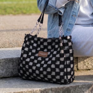 May include: A black and white checkered tote bag with silver chain straps and a small brown rectangular tag. The bag has a frayed, textured appearance and is held by a person wearing a denim jacket.
