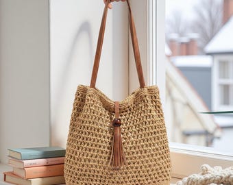May include: A woven beige tote bag with brown leather handles and a tassel. The bag is held up by a hand and sits on a windowsill next to a stack of books. The bag has a decorative wooden bead.