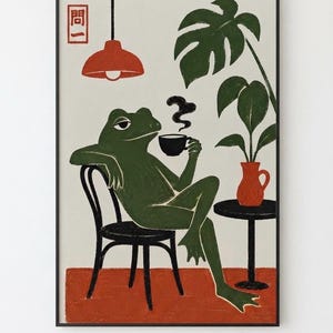 May include: A framed art print featuring a green frog sitting on a black chair, holding a black cup with rising steam. Beside the frog is a small black table with a red pitcher and a green monstera plant. A red lamp hangs above, and the Japanese text 問一 is in the corner.