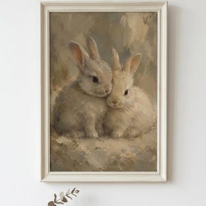 May include: A framed oil painting featuring two fluffy rabbits sitting side by side in a soft, neutral-toned setting. The artwork uses visible brushstrokes in shades of beige, cream, and light brown, enclosed in a simple, light-colored wooden frame.