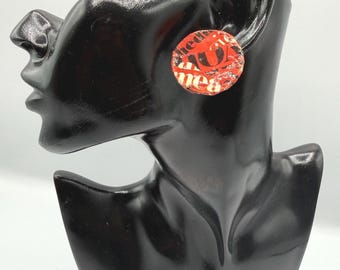May include: A circular stud earring featuring a collage of red, black, and white paper fragments with visible text including the letters U, A, and the word mea. The earring has a textured, layered appearance with a glossy finish on the surface.