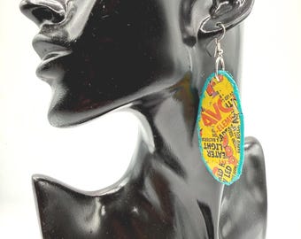 May include: An oval-shaped dangle earring featuring a yellow background with red and black printed text including the letters AVG and the word HEATER. The earring has a bright turquoise blue border and is attached to a silver-toned metal hook.